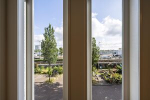 Uitzicht door hoge ramen op De Ruijterkade met bomen, geparkeerde fietsen en een moestuin, met verkeer en spoor op de achtergrond.
